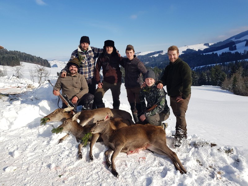 Eine Jagdgruppe mit 6 jungen Jägern steht in der verschneiten Landschaft, davor drei erlegte Tiere.